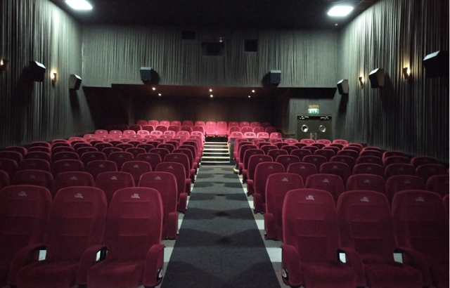 Empty theatre, facing the back. Room has middle walk way with grey carpet and red chairs either side